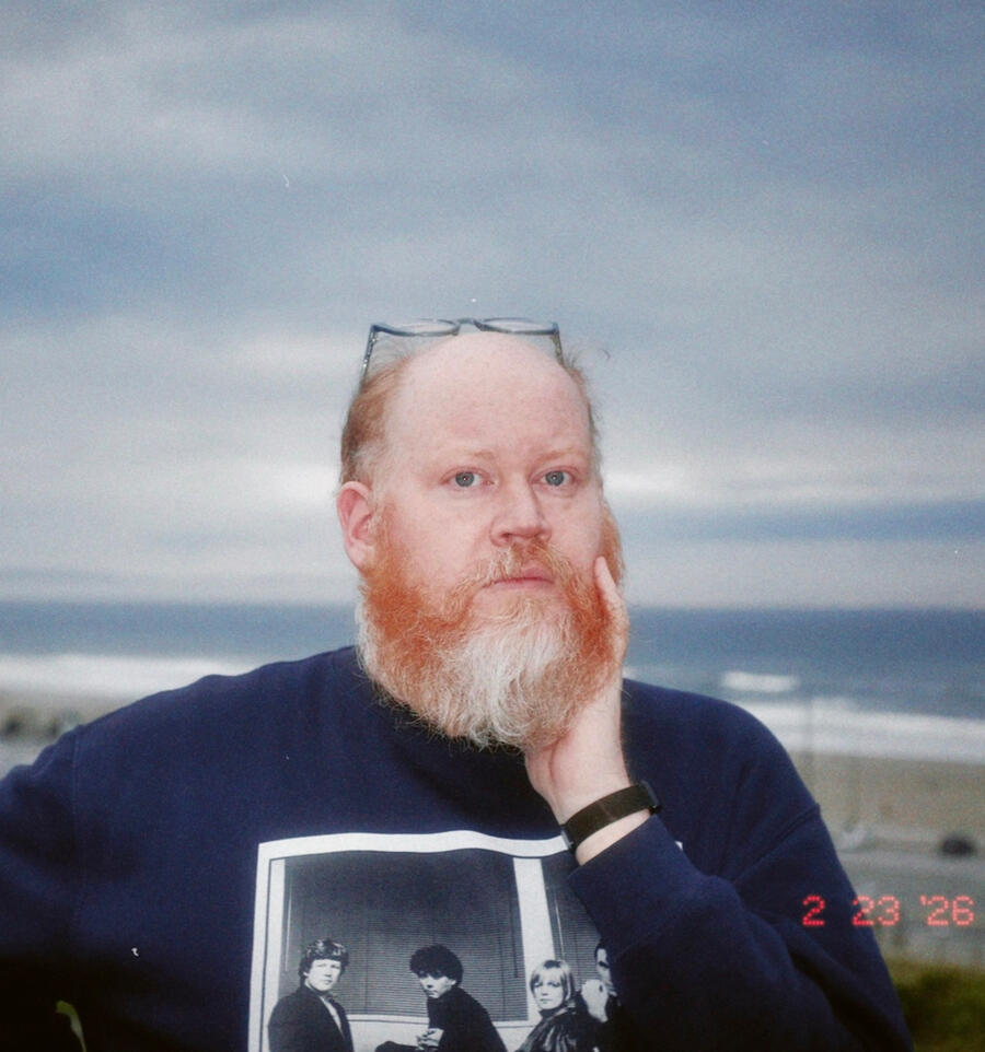 Adam Wilson, a 40-something male with balding red hair, a beard, and glasses pushed up on top of his head, wearing a blue sweater, stands in the foreground with clouds, a beach, and an ocean in the background. In the lower right corner is a date stamp show